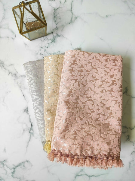 Three Amara Luxury Prayer Mats in pastel shades of silver, gold, and pink, featuring intricate floral patterns and delicate fringed edges, laid out on a marble surface beside a decorative lantern.